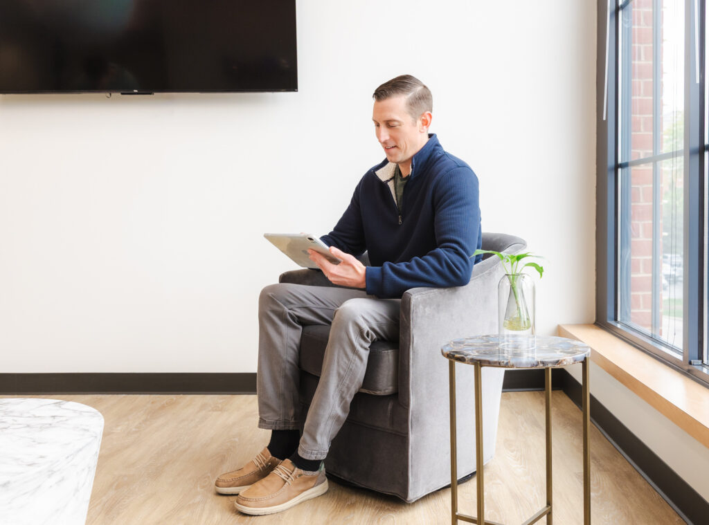 A man sitting in an armchair in a sleek, comfortable room, while holding a digital tablet. He's having a virtual conference with a Blush Aesthetics provider about weight loss injectables in Columbus.