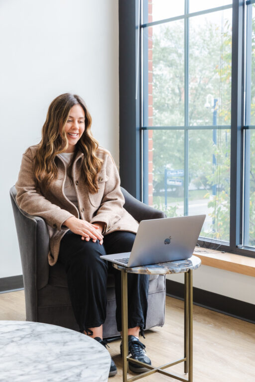 A woman sitting in an armchair looking at an open laptop on an end table in front of her.