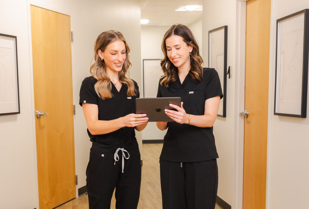 Two Blush Aesthetics team members wearing dark scrubs confer while looking at a computer tablet. They're discussing the treatment plan for a patient's lichen sclerosus treatment in Ohio.