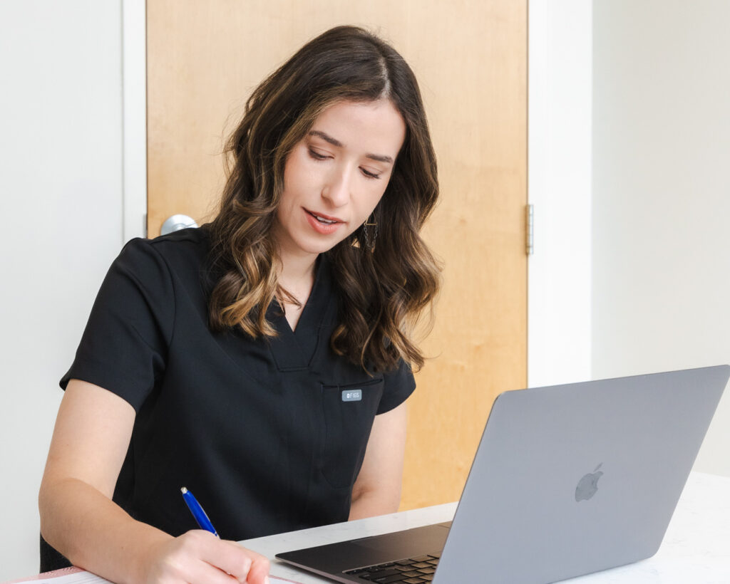 A female Blush Aesthetics practitioner, wearing black scrubs, writes down a patient's aesthetic goals and concerns before their appointment for Brazilian laser hair reduction in Columbus.